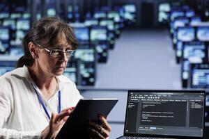 Old computer scientist writing script code in data center on laptop terminal. Adept professional doing maintenance work in server room, updating rigs software to prevent malfunctions photo