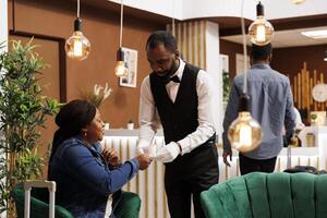 Smiling friendly African American guy waiter holding cup of fresh brewed coffee, serving it to happy female hotel guest waiting for check-in at lobby. Caffeine, travel and jet lag concept photo