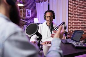 Interesting guest on online podcast show telling cool story to host, entertaining viewers watching during live broadcast. Presenter using audio recording equipment in living room studio photo