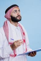 Thoughtful man wearing muslim traditional clothes looking upwards while writing in clipboard. Arab person dressed in islamic headscarf and thobe holding pen and taking notes in checklist photo