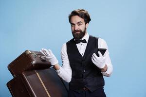 Professional bellman holds smartphone in hand and popsing next to trolley bags, portraying luxurious hospitality industry occupation. Young man in formal attire using mobile phone webpages. photo