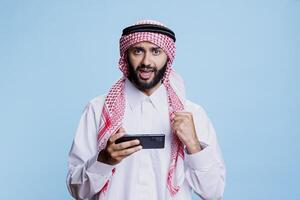 Excited arab in traditional clothes winning game on smartphone and looking at camera with clenched fist. Muslim person showing victory concept while playing videogame on mobile phone portrait photo