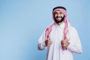 Smiling muslim man dressed in traditional clothes showing thumbs up gesture studio portrait. Cheerful arab person conveying approval and positivity sign while looking at camera photo