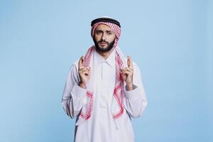 Man wearing cultural muslim clothes crossing fingers with superstitious belief and hopeful expression. Arab dressed in traditional thobe and ghutra standing with making wish and good luck gesture photo