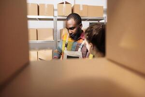 Stockroom manager explaining merchandise logistics to employee, discussing inventory report in storage room. Diverse coworkers working at customers orders, preparing packages for delivery photo
