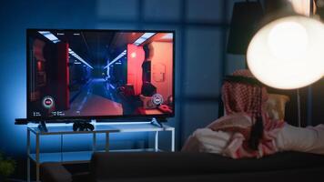 Man seeing videogame gameplay footage uploaded online by pro streamer. Arab gamer sitting at home enjoying time off from work looking up game info before deciding to buy it photo