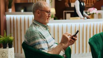Retired man checking smartphone app in lounge area at five star hotel, using online browser to navigate on social media. Senior traveller relaxing in holiday retreat lobby. Handheld shot. photo