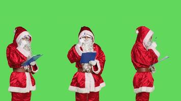 Adult in saint nick costume using papers on clipboard to create naughty and nice list, christmas eve concept. Santa claus checking to see gifts and presents, standing on greenscreen backdrop. photo