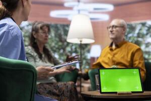 Elderly man and woman talking to nurse at their home with a green screen on tablet, having a fun chat together and asking for guidance. Senior couple feeling good, relaxing at checkup visit. photo
