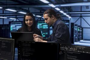 Programmers in data center using laptop to check for server performance issues, talking about solutions. Colleagues in server hub workplace fixing insufficient memory errors slowing down hardware photo
