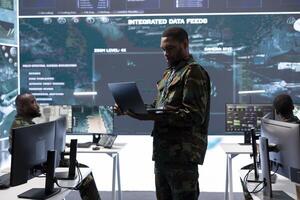 Army professional examining live satellite imagery on laptop and big screen, overseeing intelligence gathering activity in a control center. Supporting strategic military operation at the main base. photo