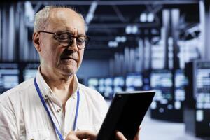 Elderly manager inspects operational server racks in computer network security data center, ensuring optimal performance. Adept technician monitoring energy consumption across components using tablet photo