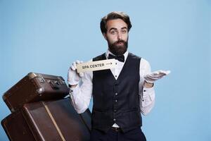 Hotel staff holds spa center pointer, guiding guests to the right direction with wall indication sign. Professional male bellhop porter posing with relaxation area indicator, blue background. photo