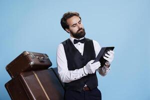 Classy porter reads guest list on device, acting like a professional and providing excellent concierge services. Bellboy with formal attire exudes sense of reliability, using tablet on camera. photo