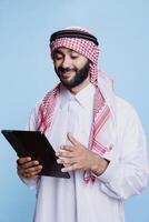 Smiling man wearing muslim thobe and headdress using digital tablet with cheerful expression. Arab person in traditional clothes scrolling through internet page on portable gadget photo