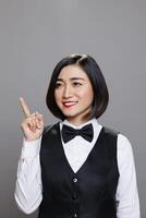 Cheerful asian woman wearing waitress uniform directing upwards with finger and posing in studio with joyful facial expression. Restaurant smiling receptionist pointing up photo