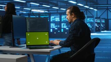 Engineer upgrading equipment in data center using green screen laptop. Specialist at PC desk in high tech establishment overseeing server rows with mockup notebook, panning shot photo