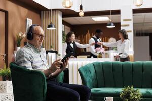 Female client checks-in at hotel reception while eldery male traveler browses and does room booking using tablet. Retired senior man with headphones, using his device for online check-in. photo