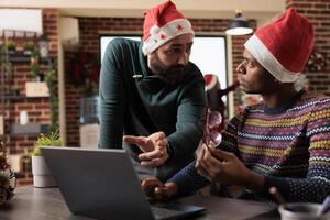 Diverse men coworkers wearing santa hats having conversation and brainstorming on laptop task in festive office. Colleagues discussing project in workplace during winter holiday photo