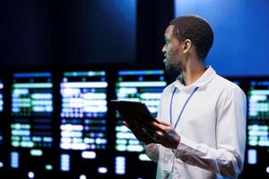 African american IT developer using tablet to audit energy consumption across server parts. Programmer making sure data center hardware temperature sensors are in flawless working condition photo