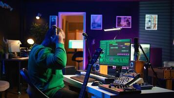 Producer composing new music on acoustic instrument in home studio, using microphone to sing chords. Musician songwriter works with mixing console and software, guitarist producing tunes. Camera B. photo