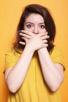 Shocked woman with her mouth covered in both hands as she stares at an unbelievable situation. Concerned female person stands against orange background, looking both surprised and terrified. photo