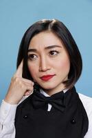 Pensive asian waitress with questioning look holding finger on head closeup. Catering service young thoughtful woman in uniform worker looking away while considering solution photo