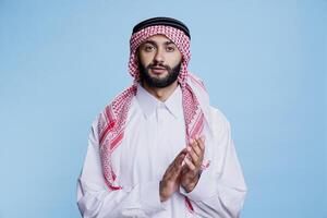 Muslim man in thobe and ghutra applauding with hands, showing support and approval studio portrait. Arab person wearing traditional outfit clapping arms and looking at camera photo