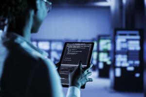 Computer operator builds firewall systems in server room housing advanced infrastructure. Facility manager runs code on tablet to upgrade data center rigs handling demanding workloads photo
