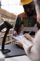 Stockroom manager signing goods inventory report, discussing merchandise quality control in warehouse. Diverse team working at customers orders, preparing packages in storehouse. Close up photo