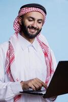 Smiling muslim man holding laptop and pressing button on keyboard with cheerful expression. Arab person wearing traditional clothes typing and sending message on computer photo