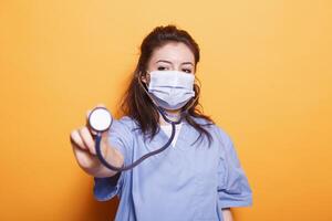 Portrait of doctor with face mask clutching a stethoscope against orange background. Nurse practitioner in the studio presenting an instrument for medical use towards the camera. photo