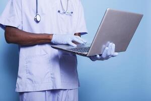 Man working as nurse using wireless computer while wearing stethoscope, gloves for protection and scrubs. African american individual standing and utilizing a digital laptop. photo