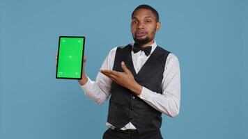 Bellman hotelier showing a tablet with green screen display in studio, presenting mockup chroma key screen against blue background. Young hotel concierge works at resort. Camera B. photo