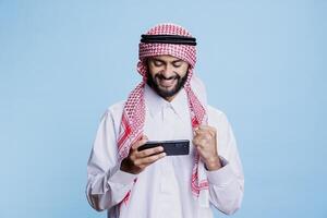 Excited man wearing muslim clothes winning mobile game and showing clenched fist in victory gesture. Arab player in traditional outfit enjoying online videogame application on smartphone photo