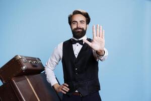Employee showing stop sign on camera, hotel concierge in suit and tie implying refusal and rejection with hand raised in studio. Professional bellboy standing against blue background. photo