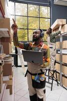 Worker relaxing listening music checking goods logistics on laptop computer, working at customer order preparing products shipment in warehouse. Manager looking at merchandise boxes photo