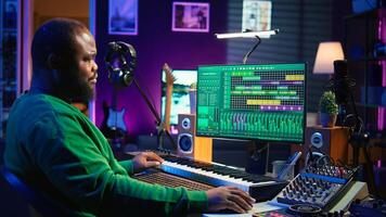 Songwriter mixing and mastering tracks on stereo console, playing electronic keyboard in home studio. Artist oversees audio performances to create melody, producing tunes with piano keys. Camera B. photo