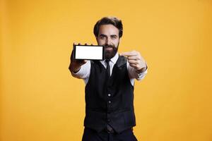 Skilled butler presents white blank display against yellow background, showing isolated mockup empty screen. Male waiter using smartphone with copyspace template in studio. photo