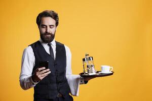Professional butler checks smartphone and preparing to serve clients at table, carrying restaurant tray with cup of coffee. Luxurious stylish employee working in catering industry, uses moobile app. photo