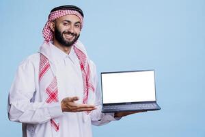 Confident cheerful man dressed in muslim clothes showing laptop with empty screen, presenting product. Smiling arab person wearing traditional attire showcasing computer blank display photo