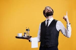Butler with food platter points up above head in studio, presenting something upwards while he prepares to serve clients at five star restaurant. Waiter employee indicates direction. photo