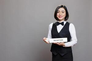 Smiling asian receptionist holding reception plate and looking at camera with cheerful face expression. Hospitality service employee in uniform showing hotel role signage portrait photo