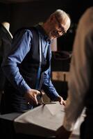 Happy couturiers in tailoring studio manufacturing premium quality sartorial garments. Senior suitmaker showing apprentice how to masterfully cut fabric and make alterations photo