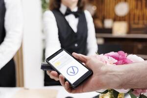 Close-up shot of caucasian old man making payment transaction using mobile phone at registration counter. Detailed view of a person using smartphone to pay for check-in reservation at hotel reception. photo
