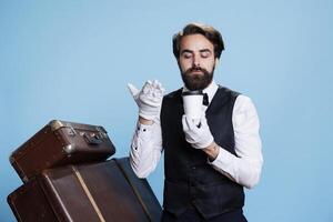 Elegant bellboy savouring smell of drink against blue background in studio, posing with cup of coffee on camera. Professional hotel concierge worker smells beverage with great aroma. photo