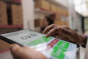 Warehouse employee checking postal form on tablet computer, searching customer parcel before start preparing order. Storehouse manager supervising cardboard boxes in storage room. Close up photo