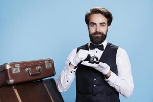 Bellboy with gloves holding service bell in studio, posing as hotel concierge employee to give assistance with luggage. Hotel worker in formal suit prepared to help guests, hospitality concept. photo