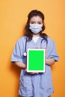 Medical doctor in hospital uniform grasps tablet showing green screen display. Nurse practitioner wearing face mask and showing blank chromakey mockup template on digital device. photo