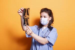 Medical practitioner with face mask analyzes radiography to find diagnosis. Assistant doctor wearing protection against virus and blue scrubs with stethoscope while looking at x ray scan in studio. photo